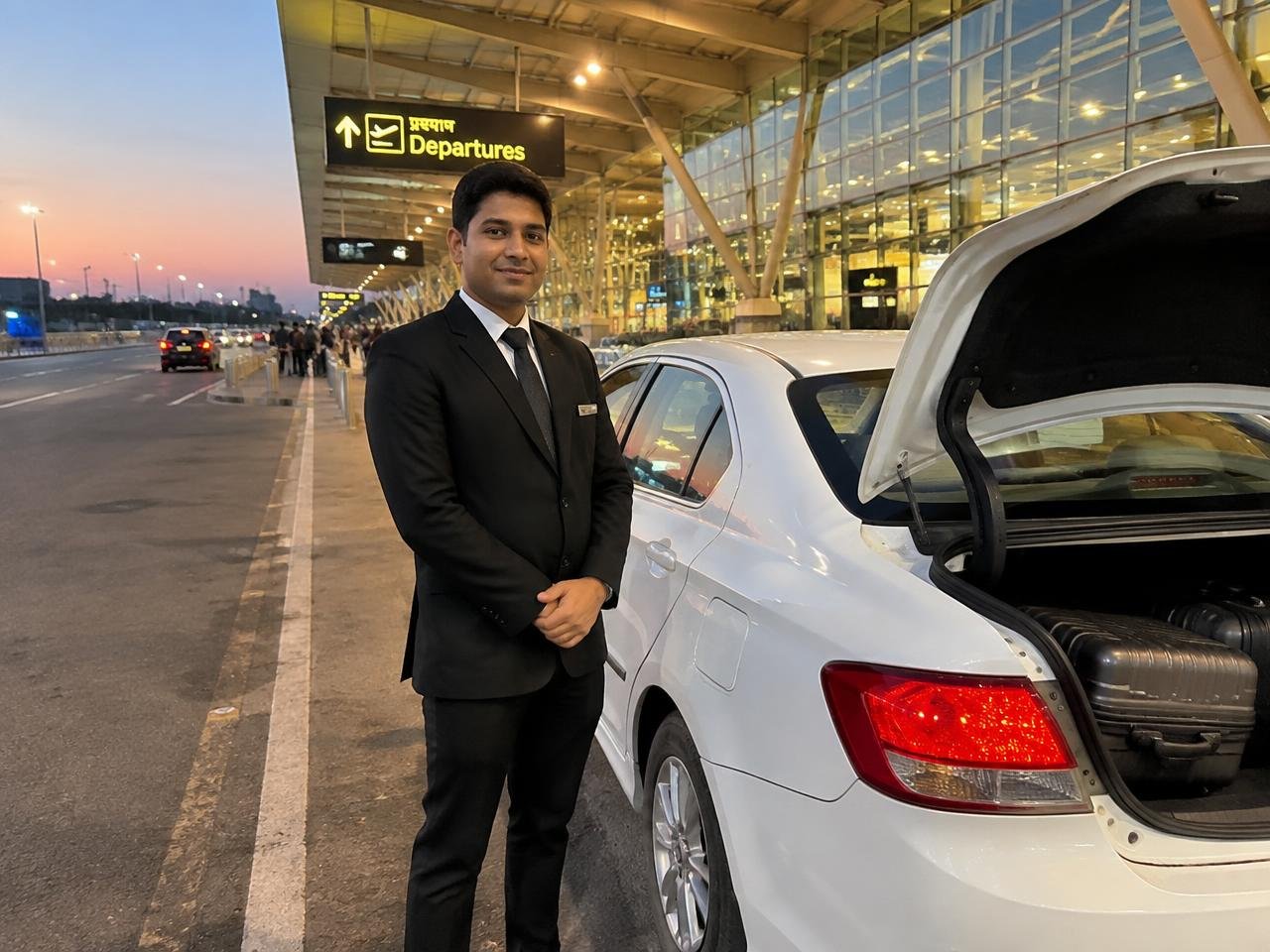 Chauffeur beside a white sedan at an airport departures terminal